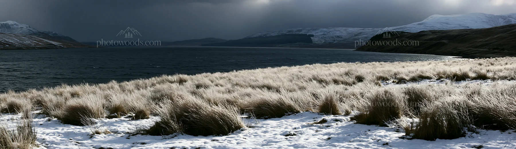 Snowy lakeside meadow with grass and mountains in the background - AI-generated stock image
