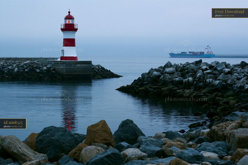 Red and white lighthouse beside a rocky harbor with cargo ship