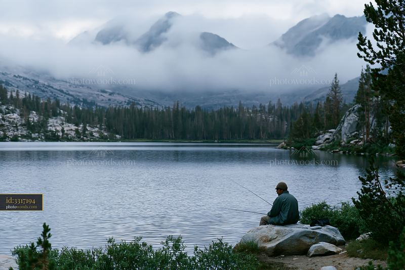 Fisherman by alpine lake beneath cloud-covered peaks