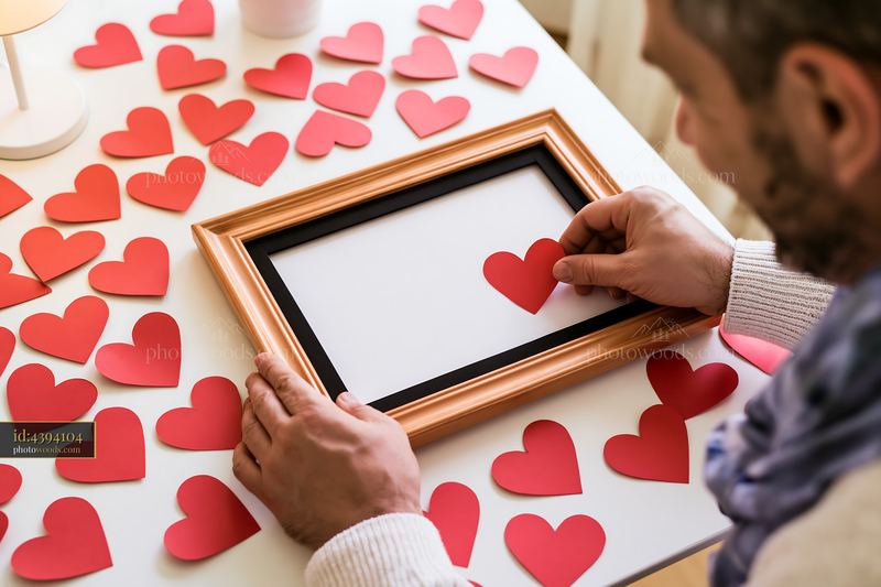 Man placing paper heart in frame on white table surrounded by hearts