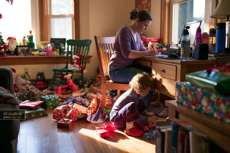 Person writing gift list at desk with child playing nearby