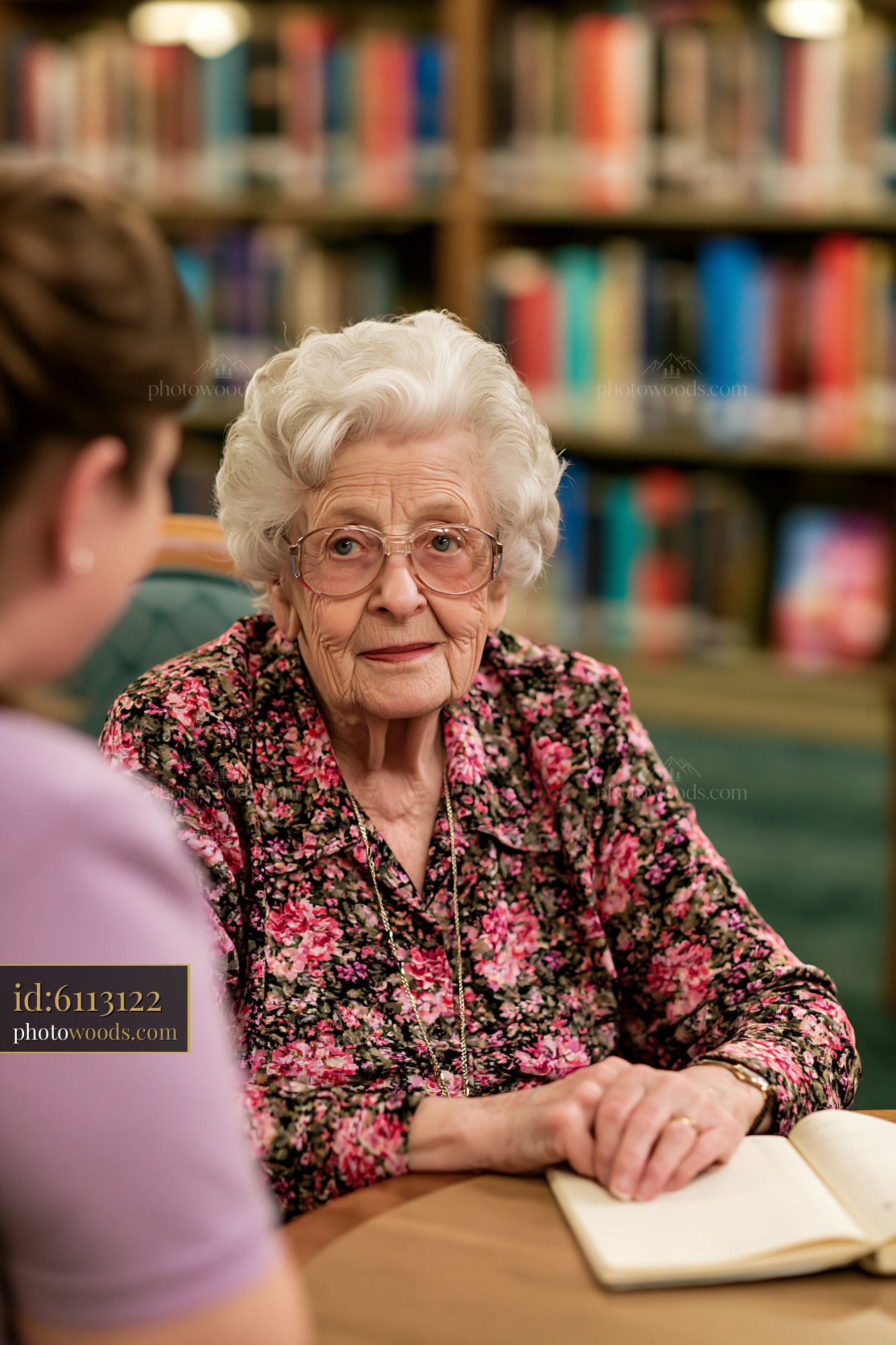 A senior woman with dementia listens attentively to a caregiver while seated at a table in a library.