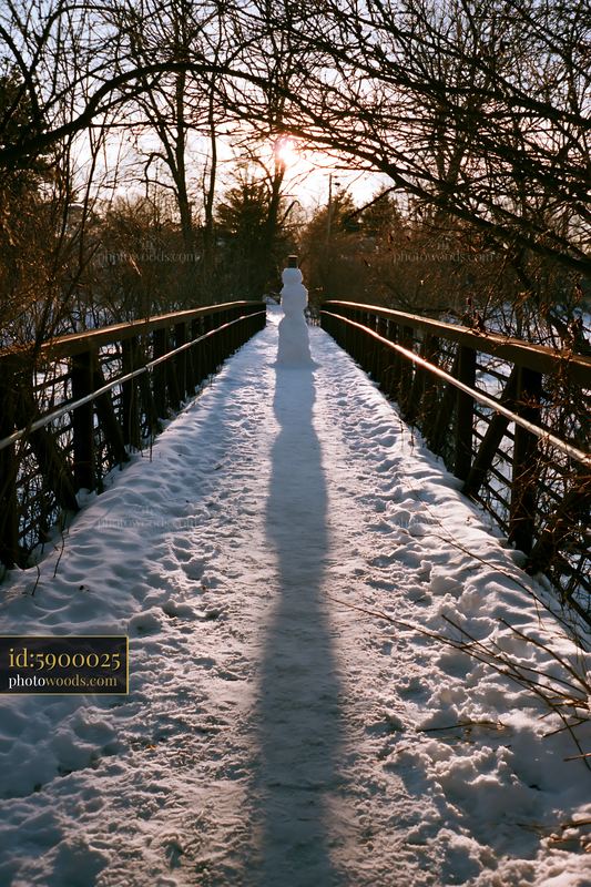 Snowman casting a long shadow on a snowy bridge at sunset
