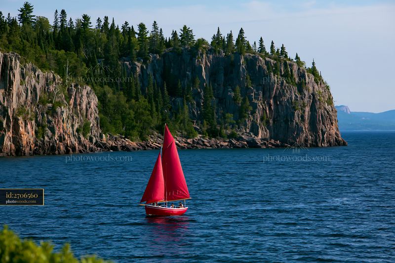 Red sailboat on a lake beneath a forested rocky cliff