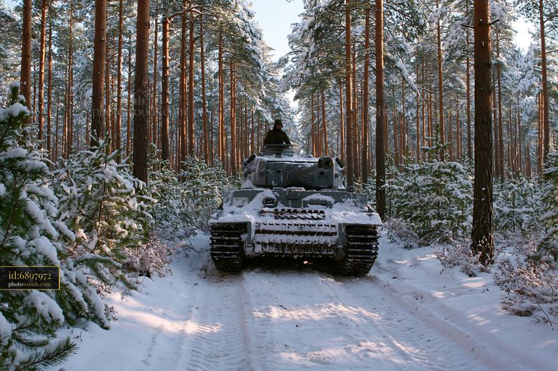 Snow-covered tank with one person in the turret on a forest trail surrounded by snowy pine trees.