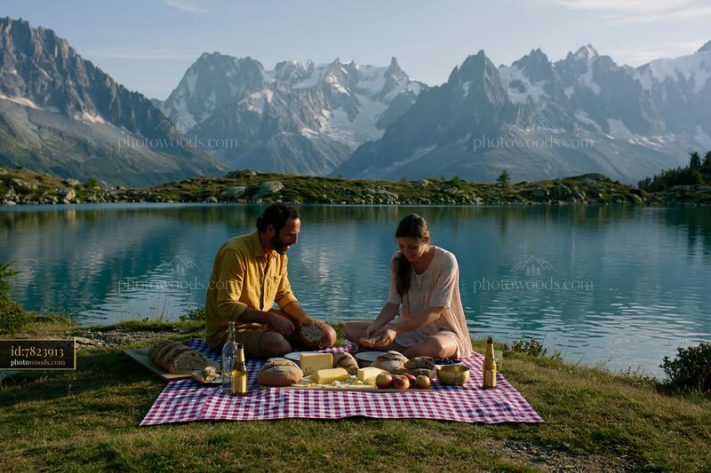 Two people share a picnic beside an alpine lake with snowy mountains in the background.