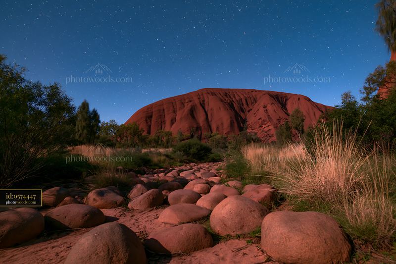 Uluru under a starry night sky with rocks in the foreground