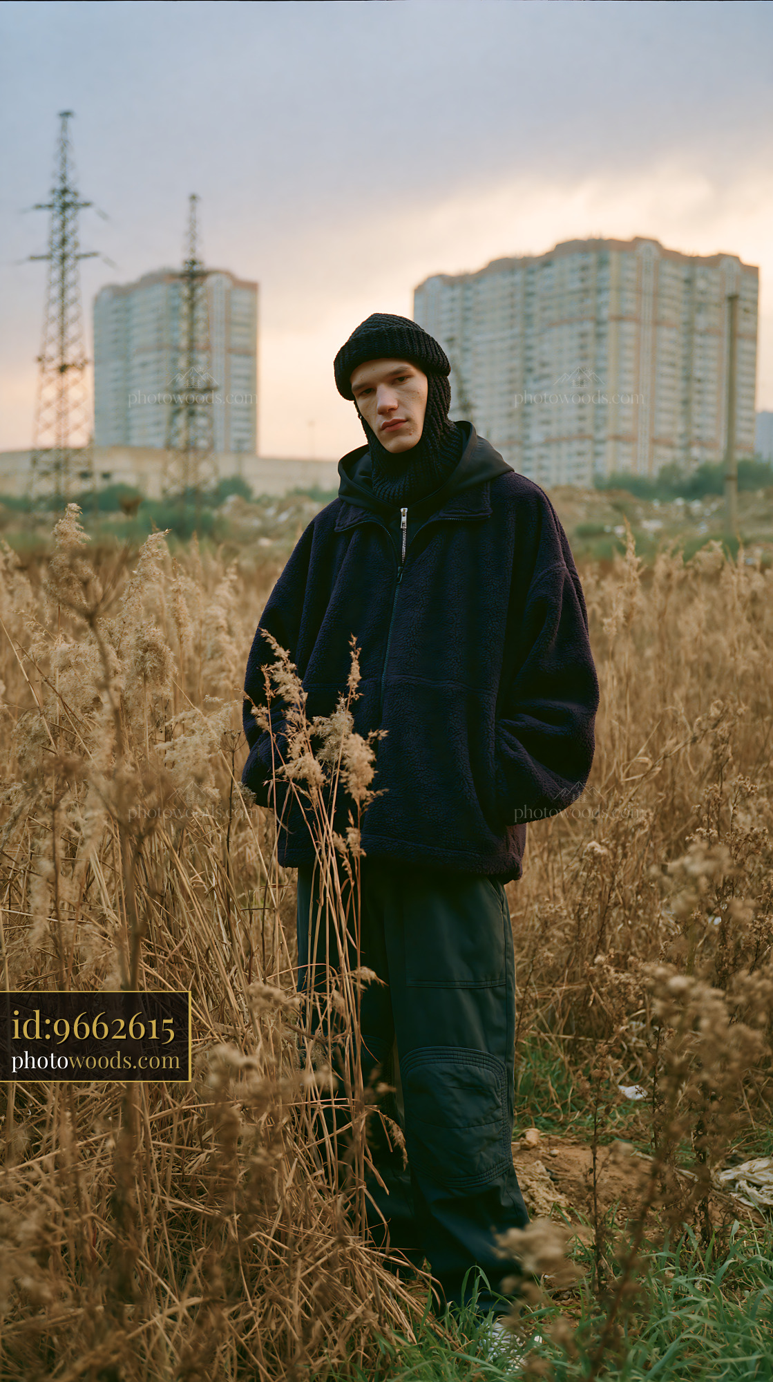 Young Man in Knitted Balaclava Stands Among Tall Grass at Sunset